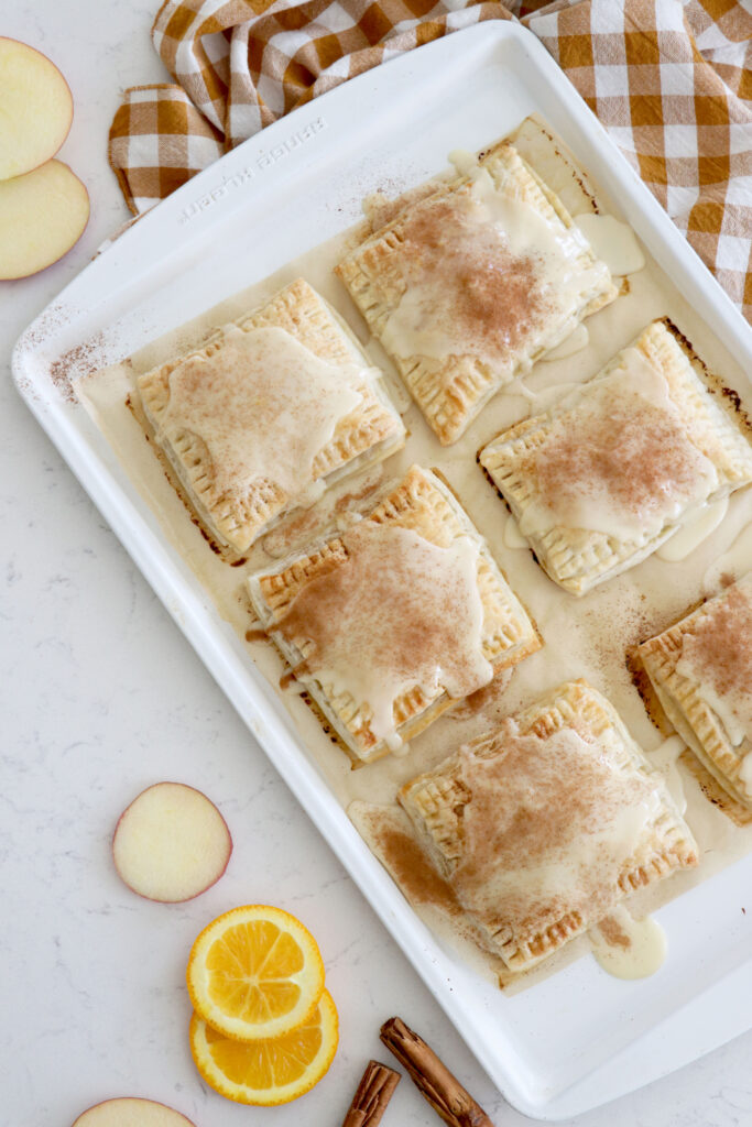 white baking sheet with apple filled pop tarts and some orange slices and cinnamon sticks on the side.