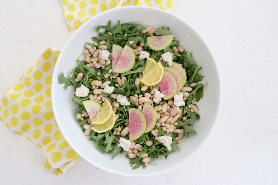 White salad bowl with lemon radish arugula salad and yellow napkin on left side.