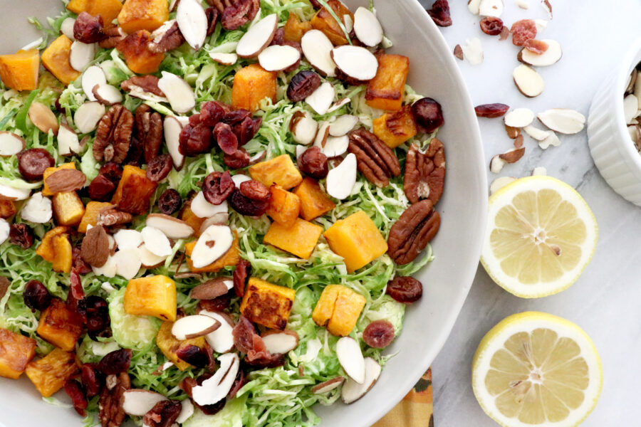 Large white bowl filled with Brussels sprouts butternut squash salad with cranberries all on a colorful napkin. Lemons and cranberries on the side.