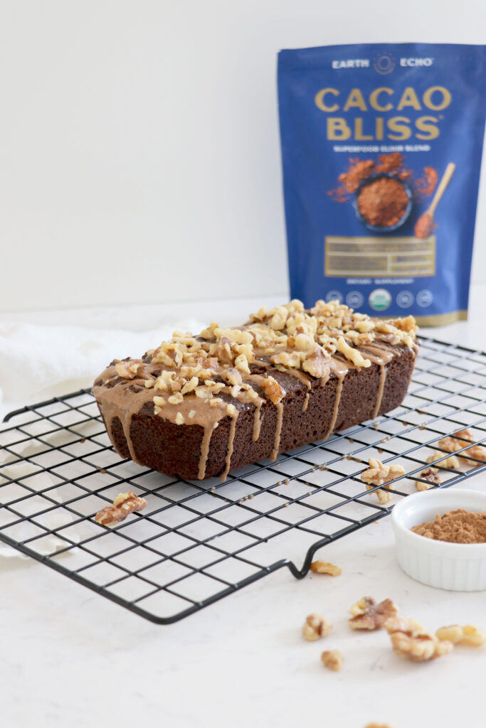 Cacao loaf topped with walnuts on wire rack with cacao bliss in background.
