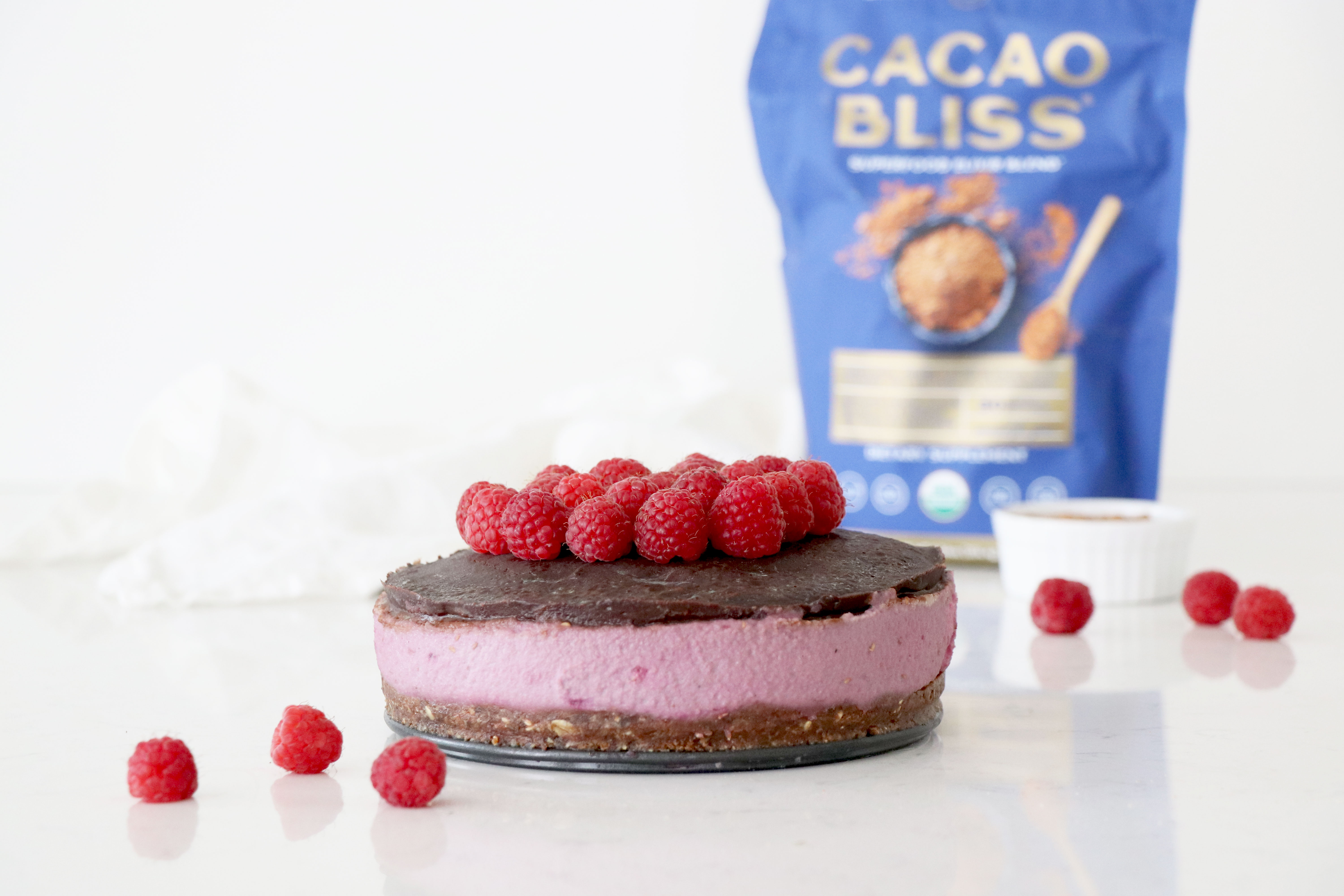 Chocolate Raspberry Cheesecake on white marble counter with Cacao Bliss package in background and raspberries on counter