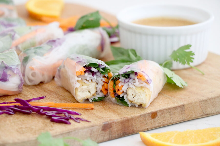 Wooden cutting board with spring rolls on top filled with veggies and a white bowl with almond pb sauce.