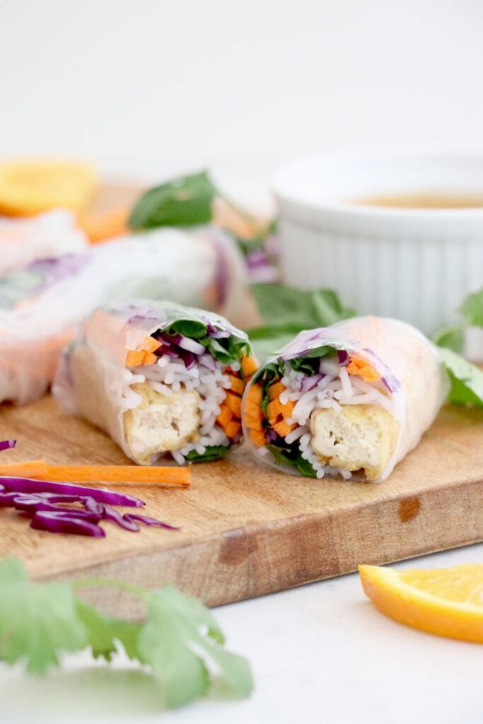 Spring roll cut In half on wooden cutting board surrounded by veggies.