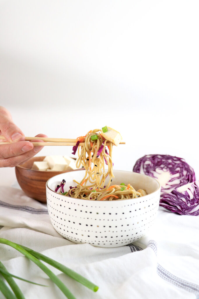 A bowl of Edamame Spaghetti and chop sticks pulling noodles up into the air