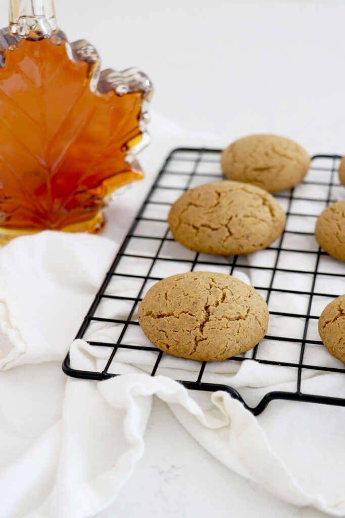 Maple syrup on top left and wire rack with ginger maple cookies all on white napkin.