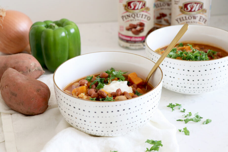 Bowls of chili with gold spoon surrounded by sweet potato, green bell pepper and beans.