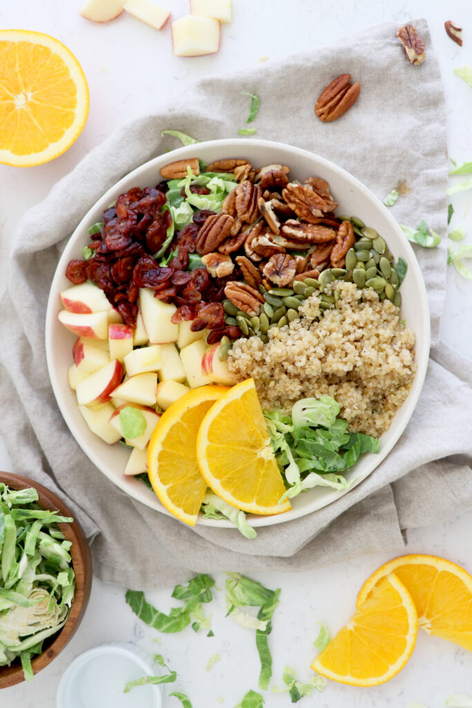 White bowl with greens, quinoa, orange slices, diced apple and pecans.