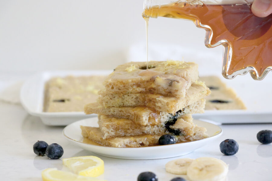 White plate with lemon blueberry pancakes and maple syrup being drizzled on top.