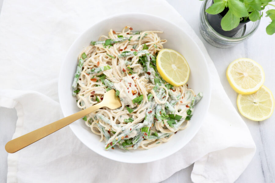 White bowl filled with pasta, cashew cream sauce and basil leaves. Gold fork in bowl and basil on the side.