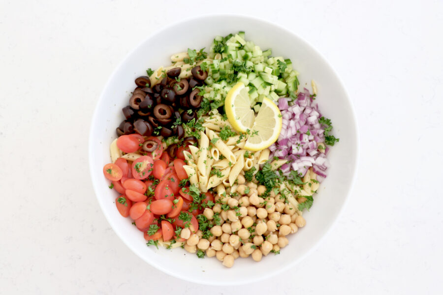 Big white bowl with pasta salad topped with diced onion, cucumber, chickpeas, olives and tomatoes.