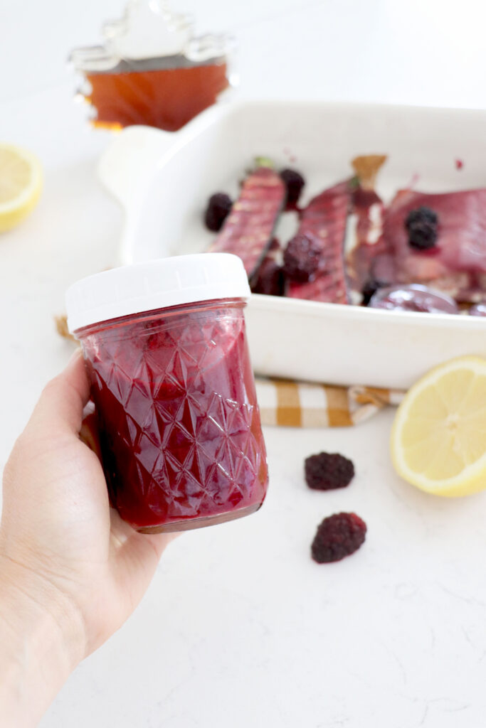 Hand holding jar of blackberry sauce with grilled food in background