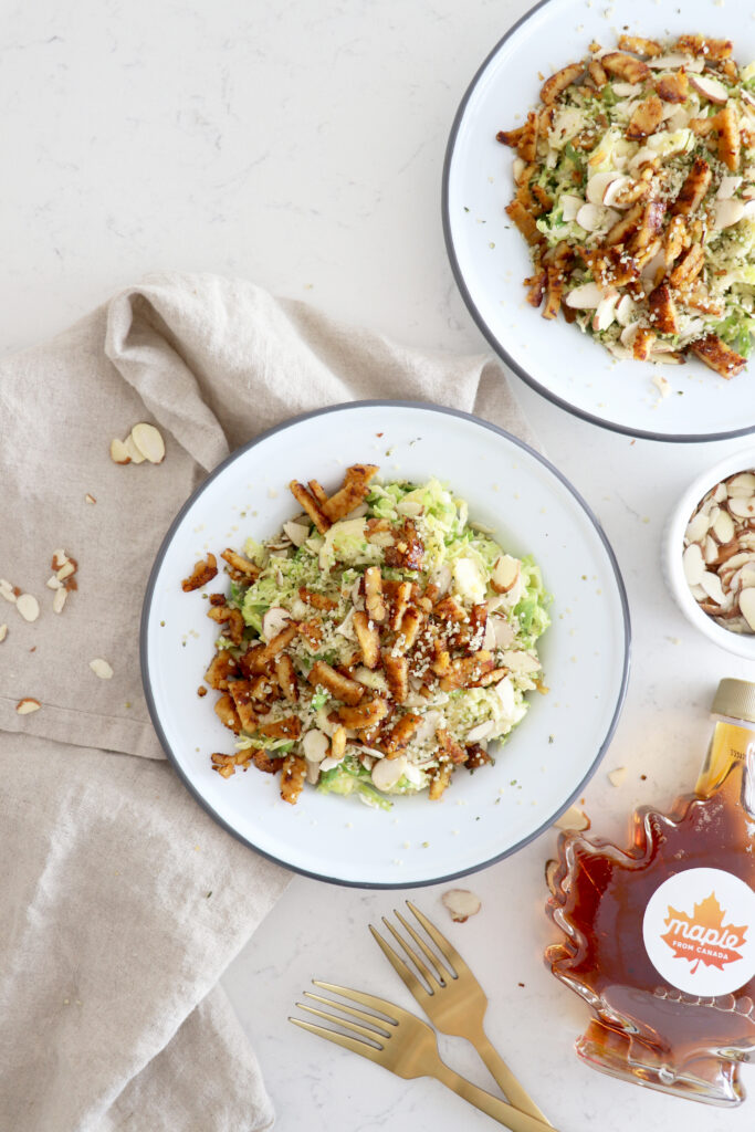 Two plates of tempeh bacon salad with maple syrup bottle off to the side.