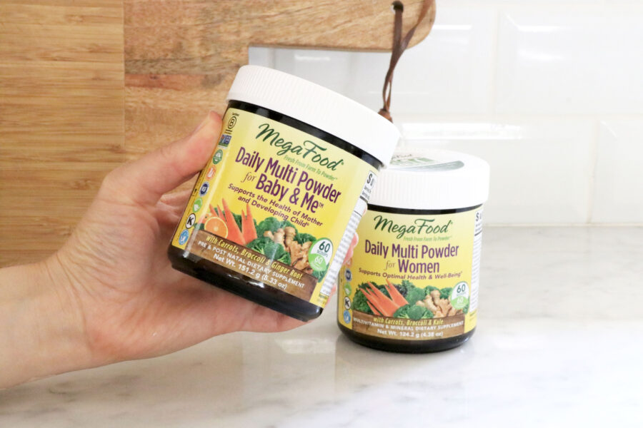 2 MegaFood multi vitamin powders in center of photo. Hand holding the left powder. Brown cutting board in background. 