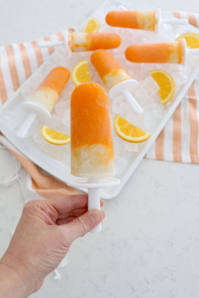 Hand holding creamsicle popsicle with other popsicles on ice behind.