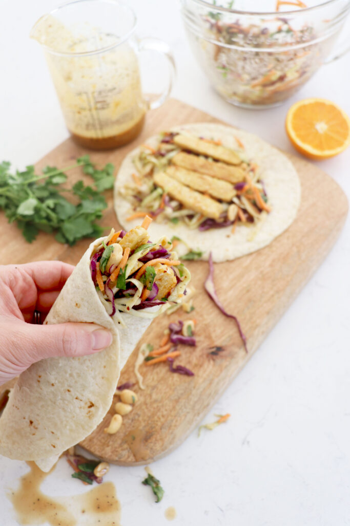 Hand holding tempeh wrap with board and wraps in the background.