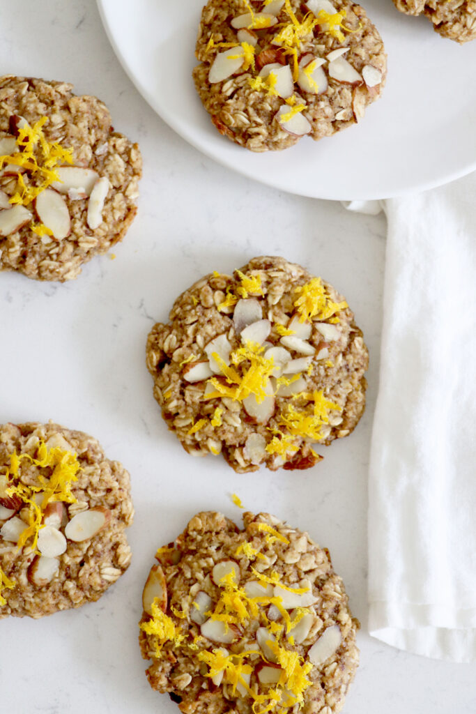 oatmeal cookies topped with almonds and orange zest on counter with plate in top right corner.