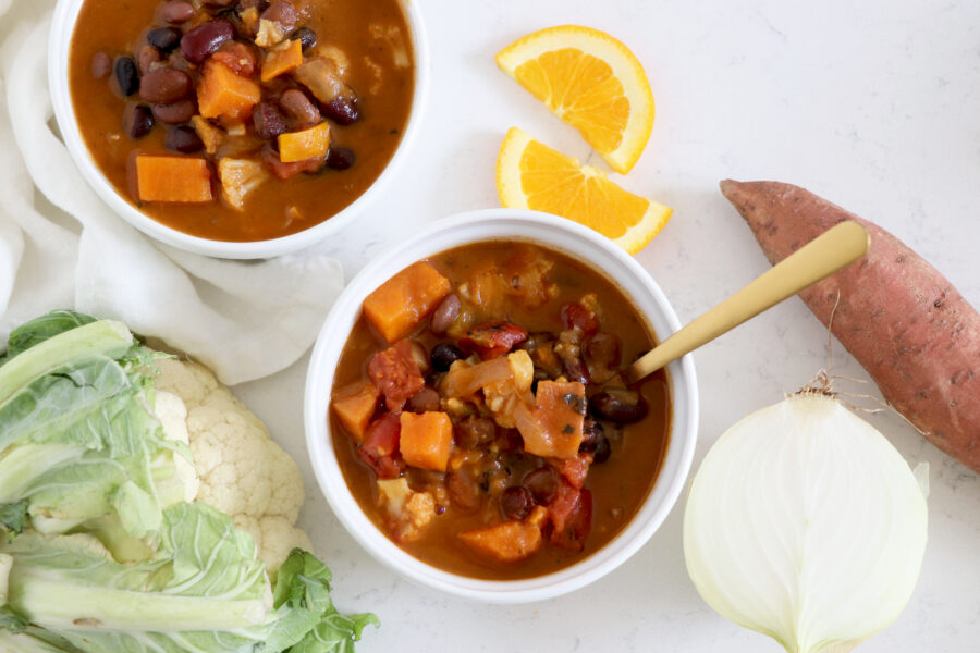 Two white bowls filled with chili surrounded by cauliflower, sweet potato, sliced onion and orange slices.