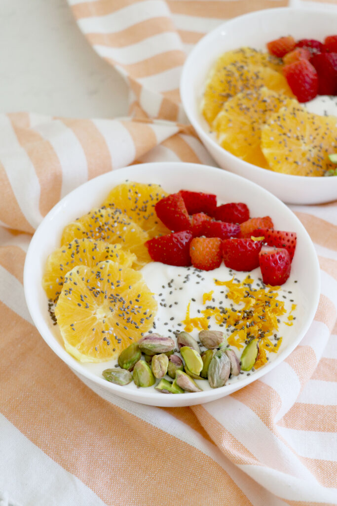Pink striped napkin with white bowls on top. Sliced fruit and pistachios in yogurt bowls.