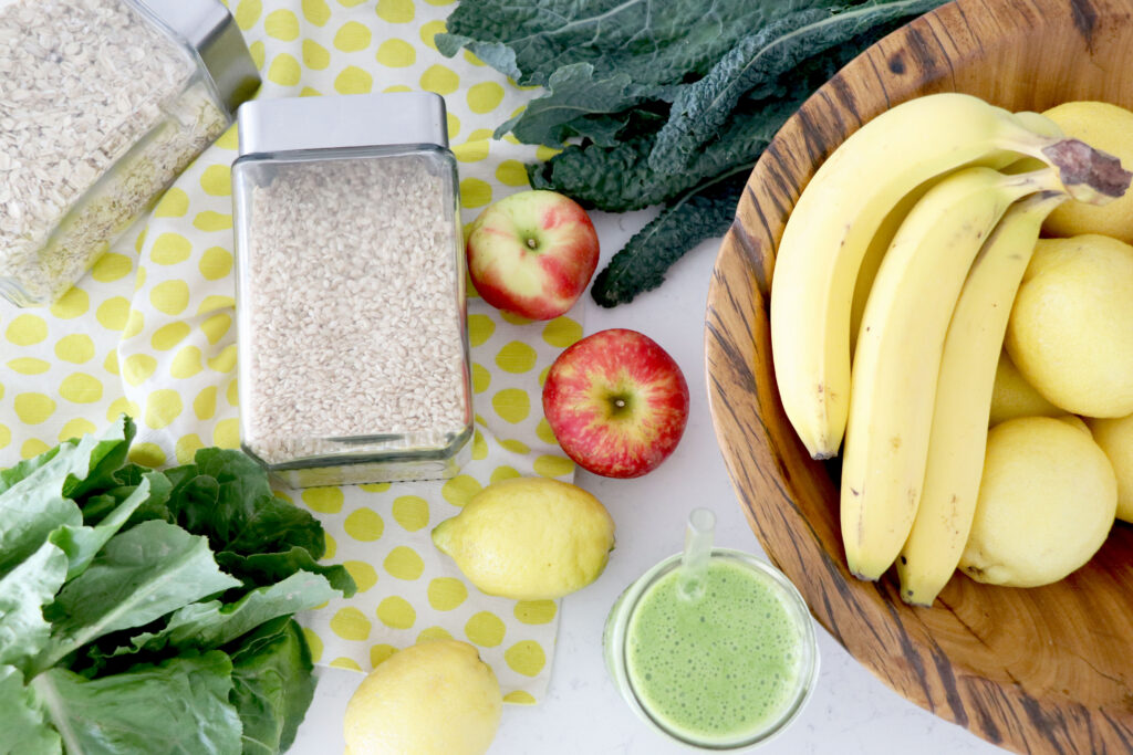 Counter with kale, greens, bananas and lemons in a brown basket, seeds in glass jars and a green drink.