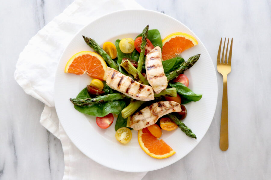 White plate of salad topped with asparagus, orange slices, tomatoes and grilled chicken. Gold fork on the right. 