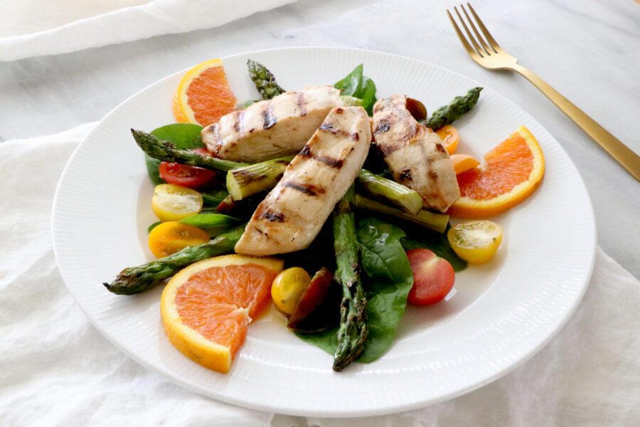 White plate of salad topped with asparagus, orange slices, tomatoes and grilled chicken. Gold fork on the right. 