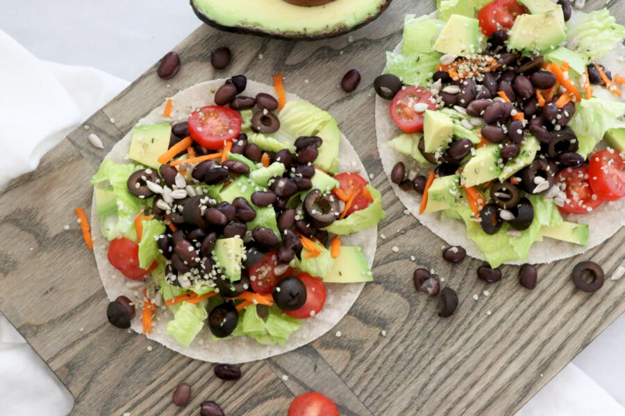 Two tacos on wooden board topped with beans and salad. Avocado half in top center.