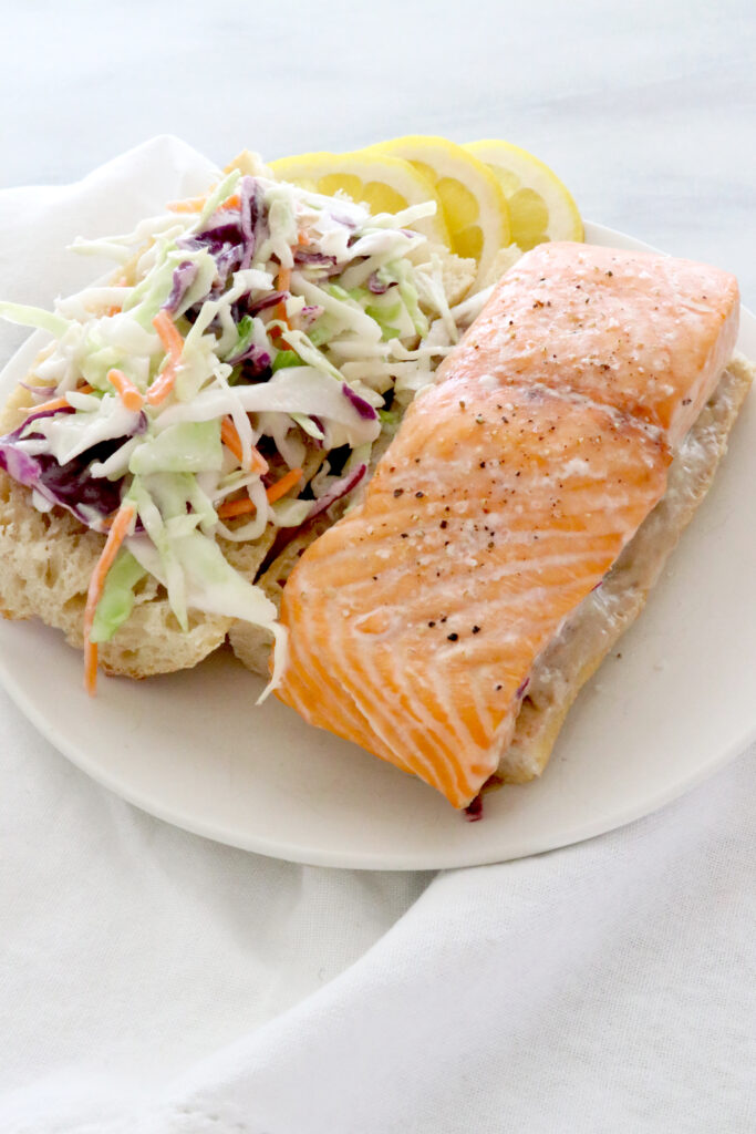 Salmon sandwich on white plate in center of photo sitting on top of a white napkin.