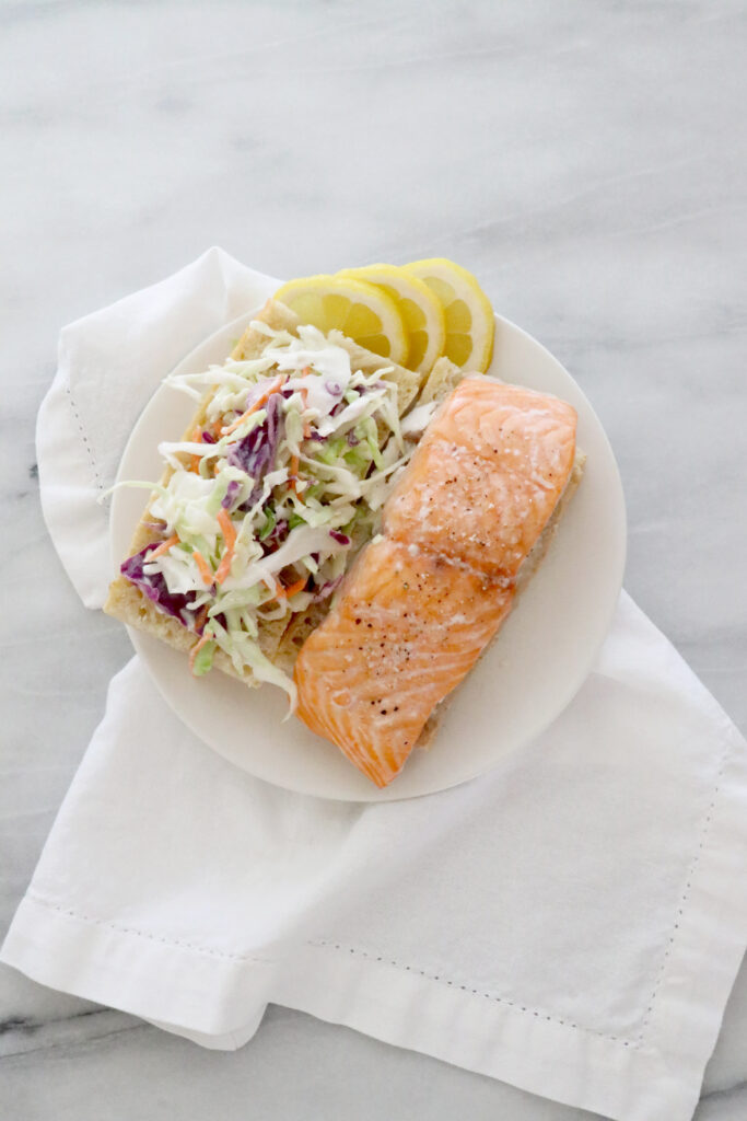 Salmon sandwich on white plate in center of photo sitting on top of a white napkin.