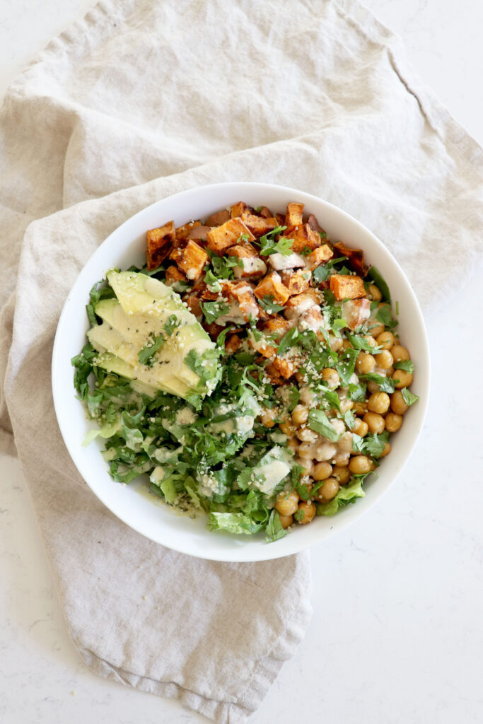 White bowl with romaine lettuce, roasted chickpeas and sweet potato with tahini dressing and avocado