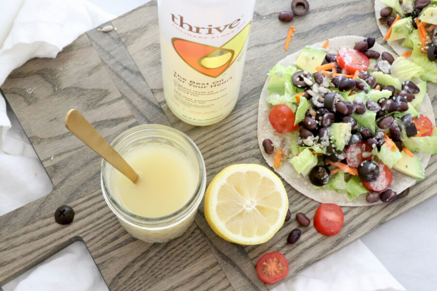Lemon Tahini Dressing in center of photo with spoon in glass container. Half of lemon next to dressing. Thrive algae oil in picture and salad taco on the side.