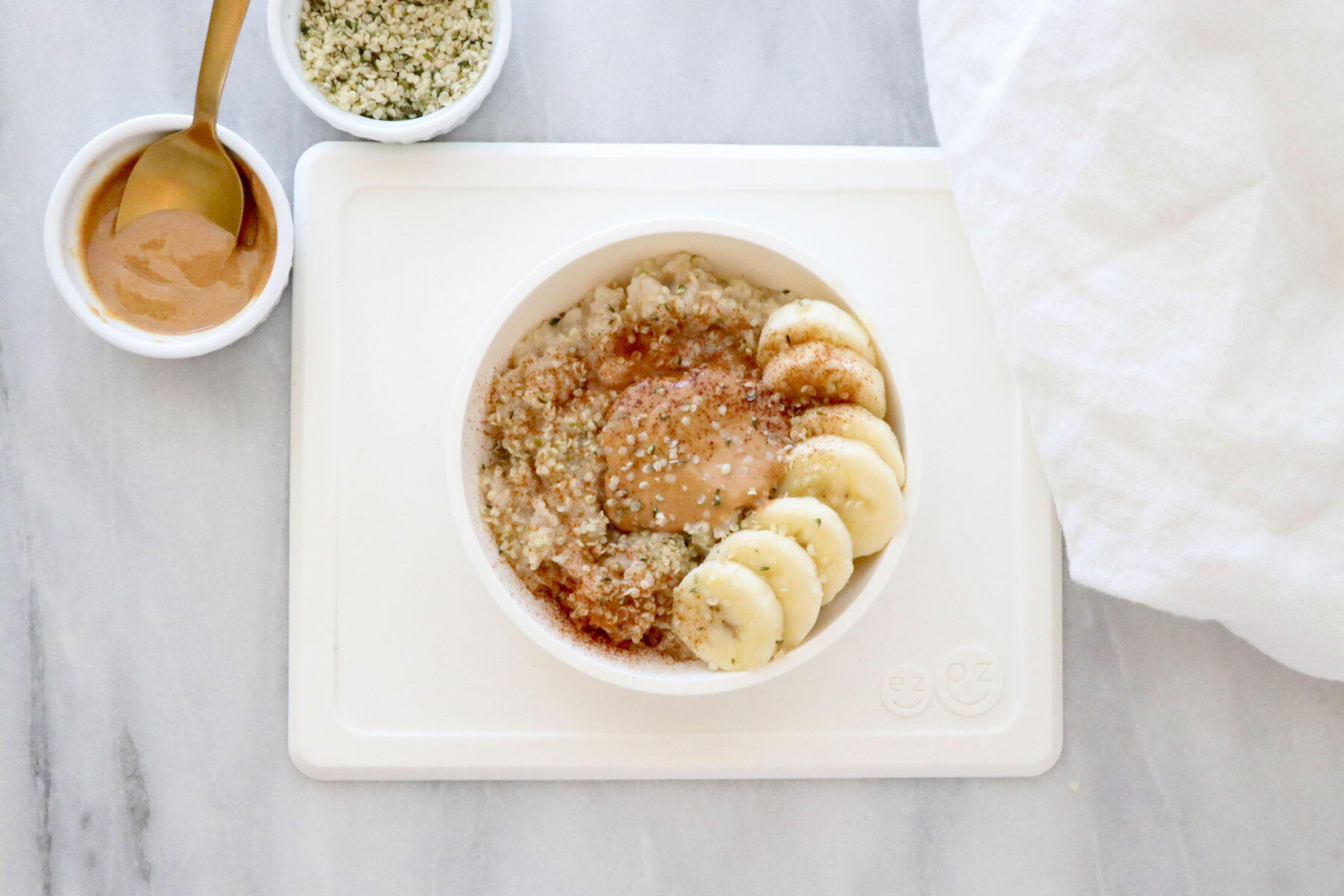 White toddler plate filled with oatmeal, topped with cinnamon, hemp seeds and sliced bananas. Small bowls of cinnamon and hemp seeds on the left side and white napkin on the right.