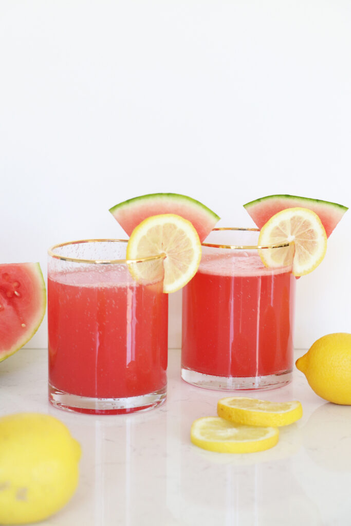 Two glasses filled with pink watermelon lemonade. Sliced watermelon and lemon on glass rim.