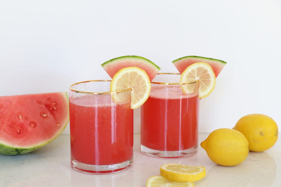 Pink watermelon lemonade in two glasses with sliced fruit on rim. Watermelon half and lemons on each side.